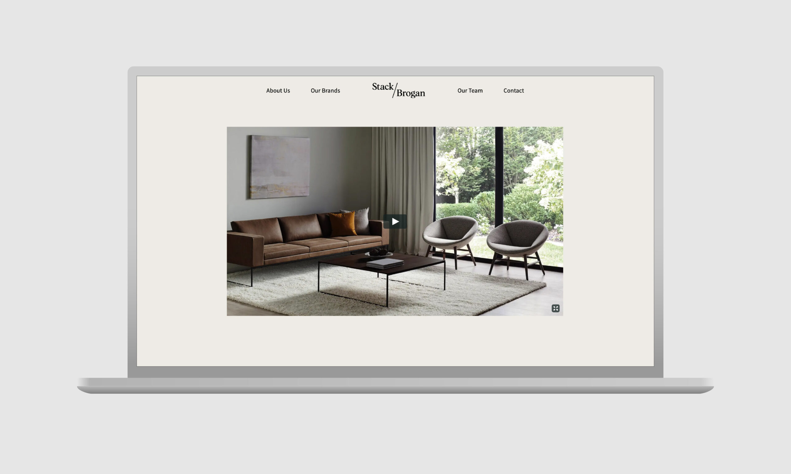 Laptop displaying the home page of the Stack Brogan website — a minimalist living room image with a brown sofa, two armchairs, and a coffee table on a rug. Large window shows greenery outside.