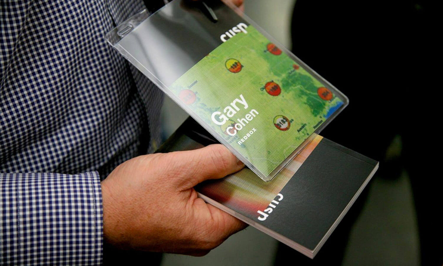 A person holds a Cusp booklet and a green name badge with the background blurred.
