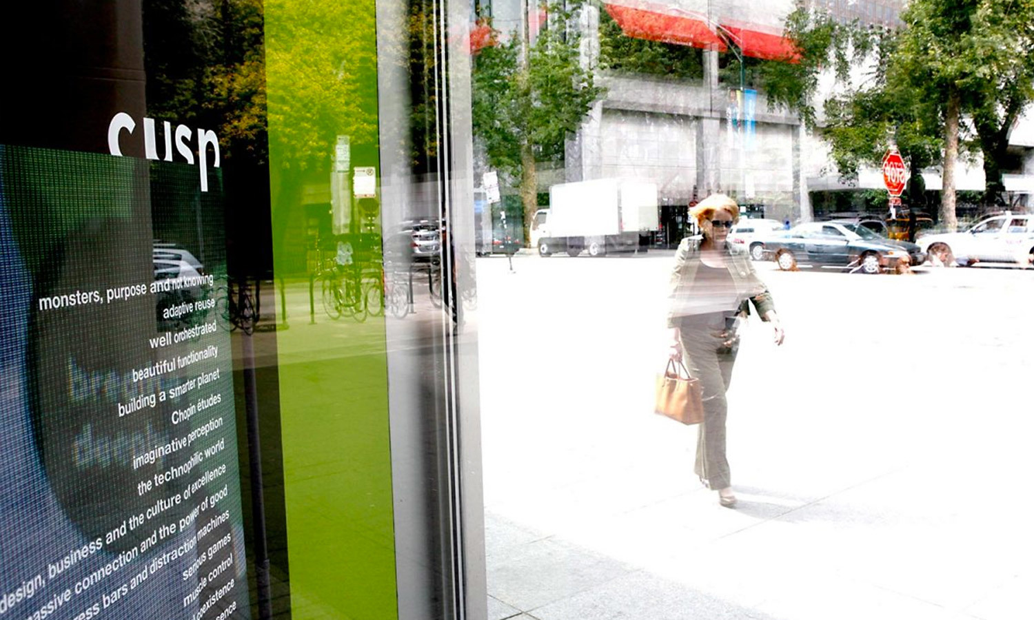 Reflection of a woman walking past a city storefront window displaying a poster about Cusp. The street outside is bright and bustling with vehicles.