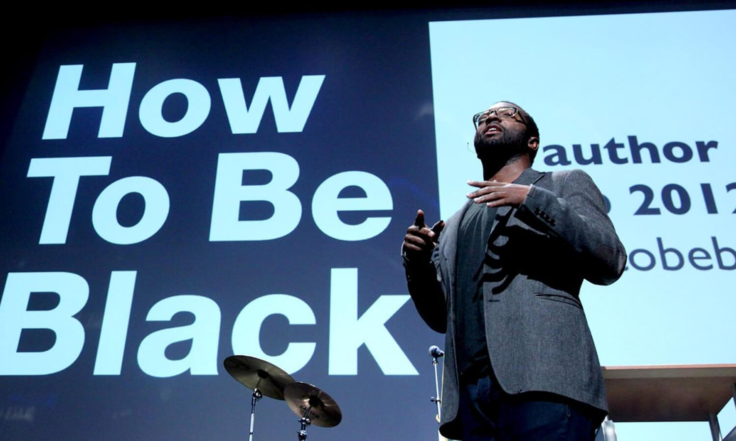 A speaker stands confidently on stage at the Cusp conference in front of a large screen displaying the text "How To Be Black," conveying a theme of empowerment and engagement.