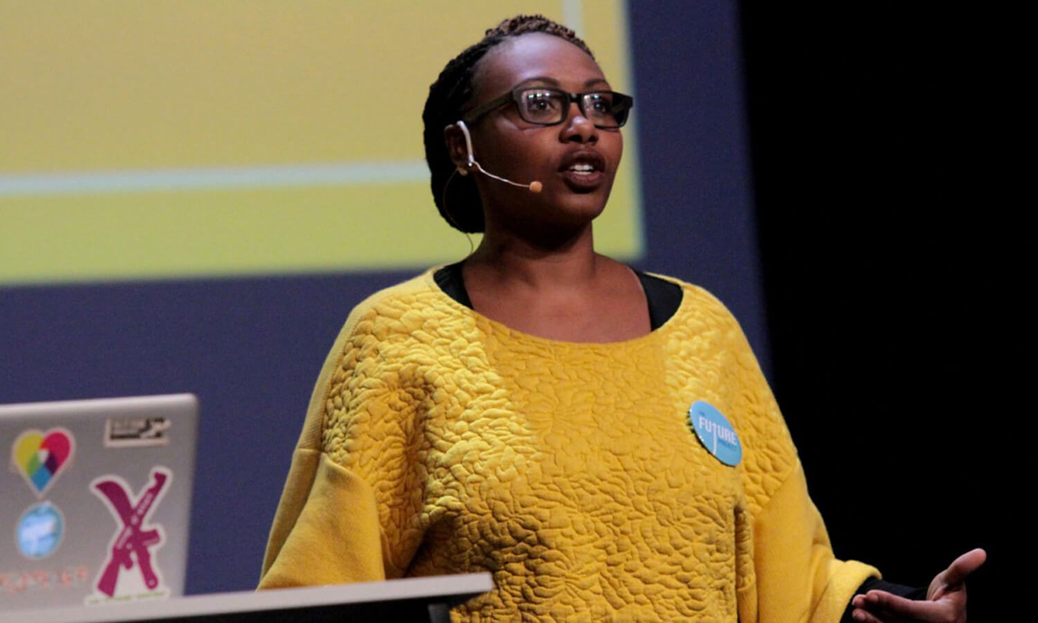 Woman in a yellow sweater speaks confidently on stage at the Cusp conference with a headset mic. She stands near a laptop covered in colorful stickers.