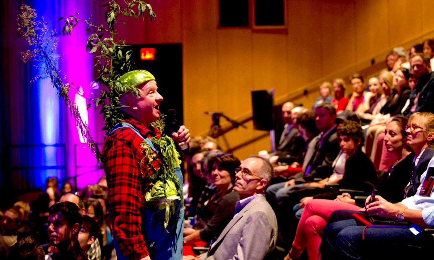 A man in a leafy costume and green hat speaks into a microphone. He stands among an amused audience in a brightly lit auditorium.