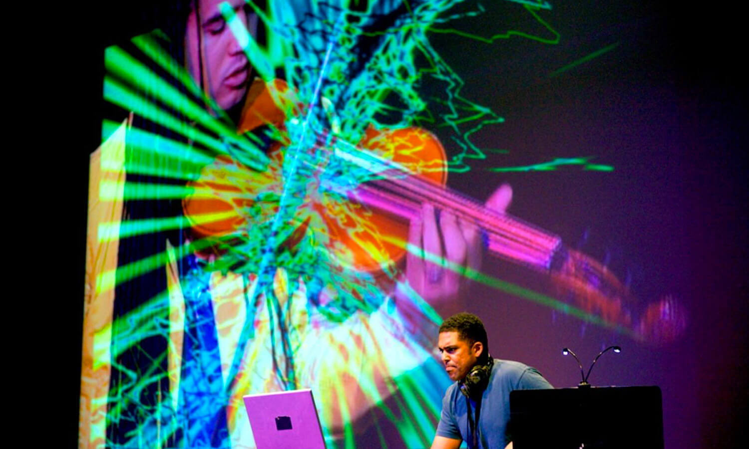 A DJ performs live at the Cusp conference, focused on his laptop, while vibrant, colorful visual projections of a violinist and abstract patterns fill the background, creating an energetic atmosphere.