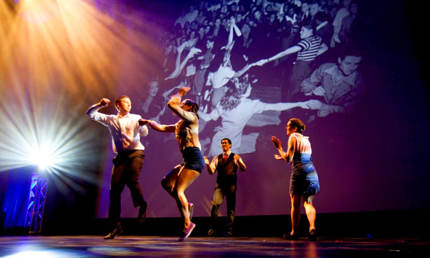 Dancers energetically perform on stage at the Cusp conference under bright lights, with a large, vintage black-and-white photo of a lively dance scene projected behind them.