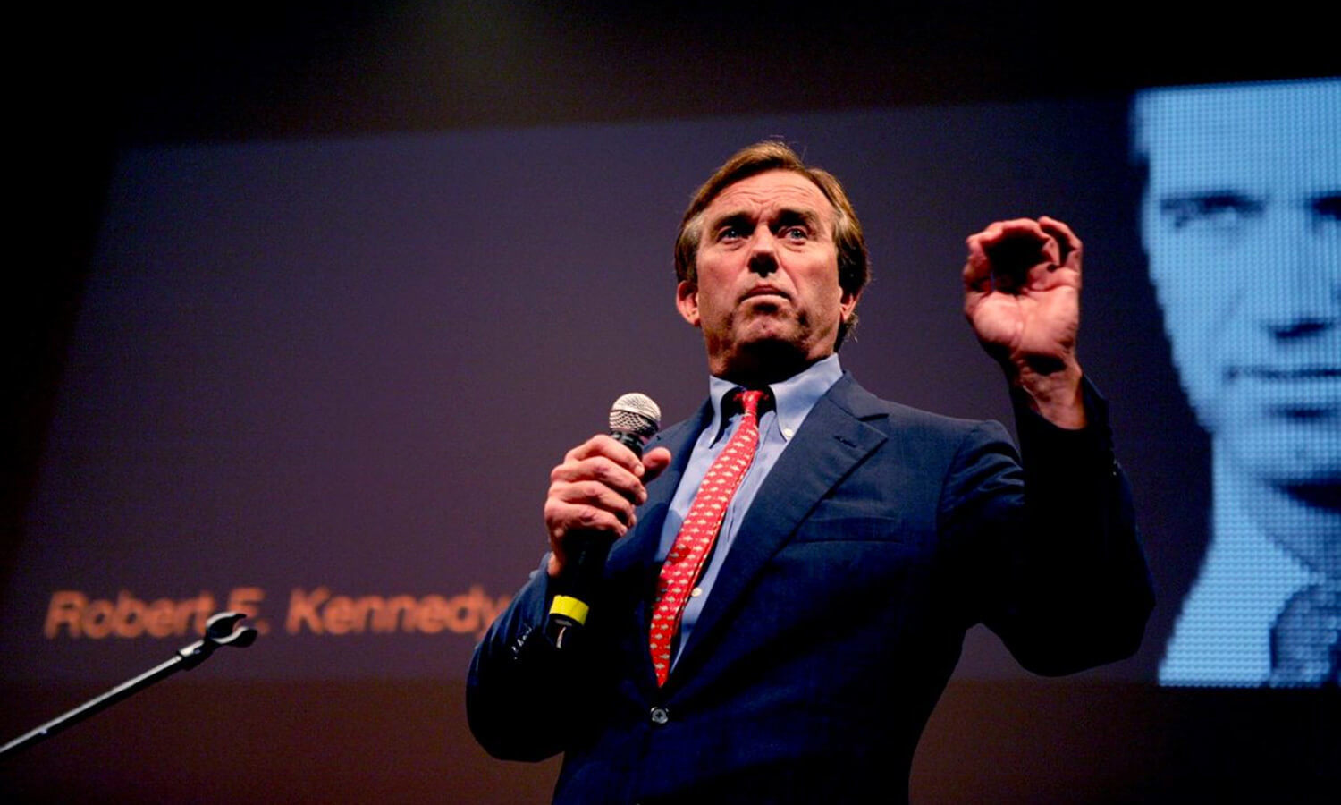 A man in a suit with a red tie speaks into a microphone on stag at the Cusp conference.