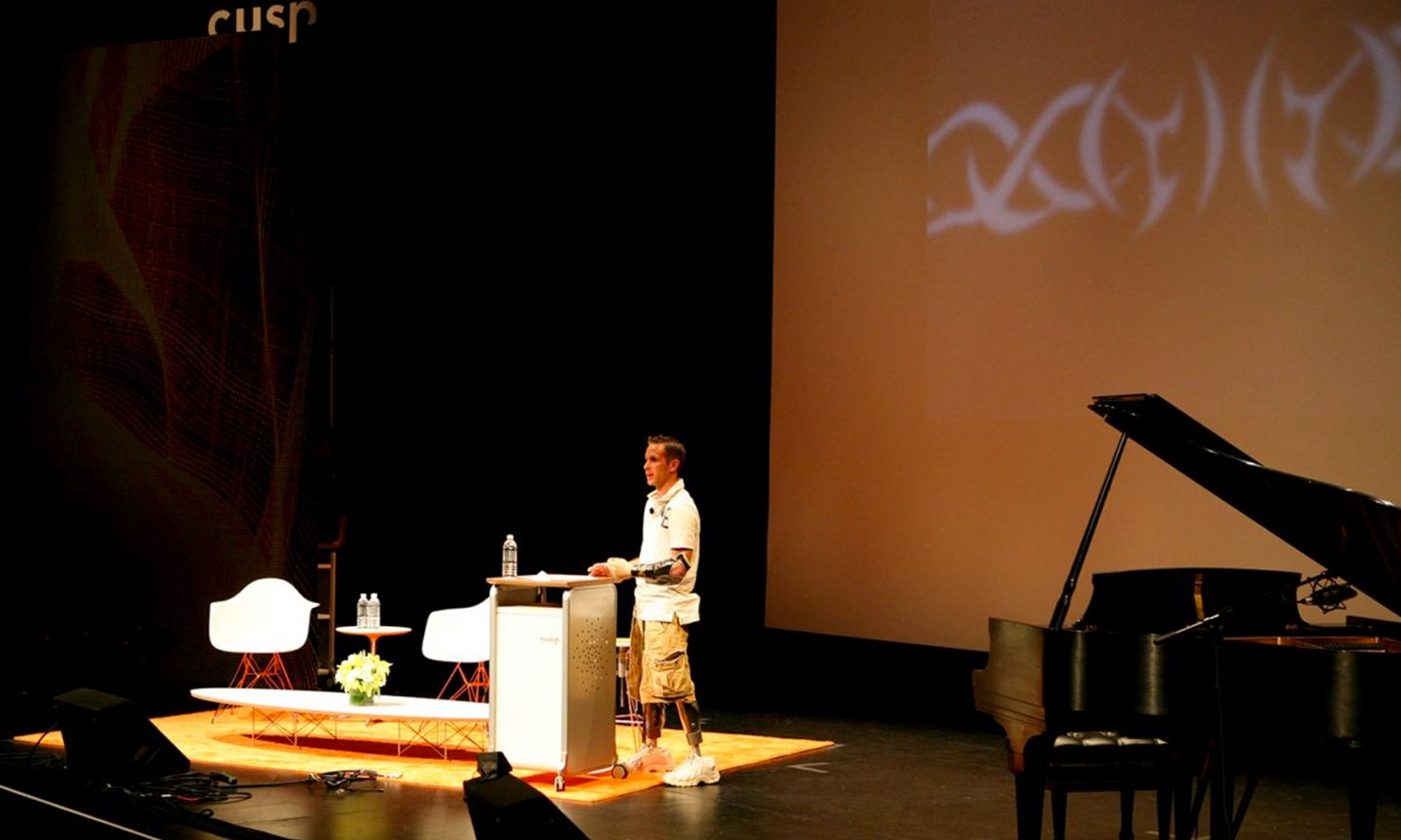 A person stands at a podium on a dimly lit stage, delivering a presentation at a Cusp conference with a screen showing abstract designs. A grand piano is visible on the right.