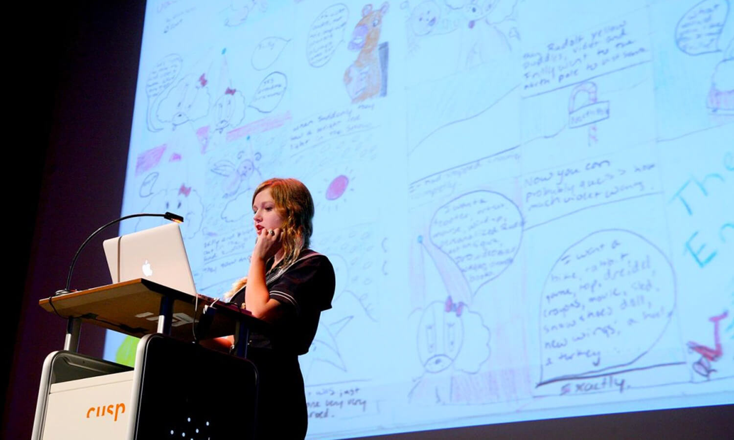 A woman stands at a podium presenting at the Cusp Conference in front of a large screen displaying colorful, hand-drawn sketches and notes.