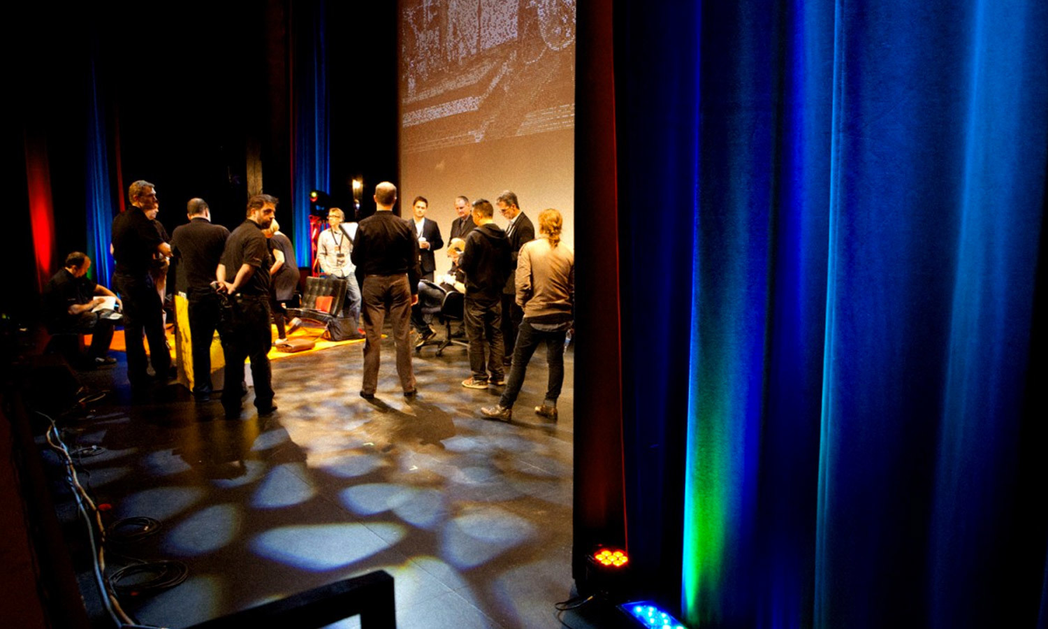 A group of people stands on a dimly lit stage in discussion about the Cusp conference, surrounded by colorful blue and red curtains and patterned floor lighting.
