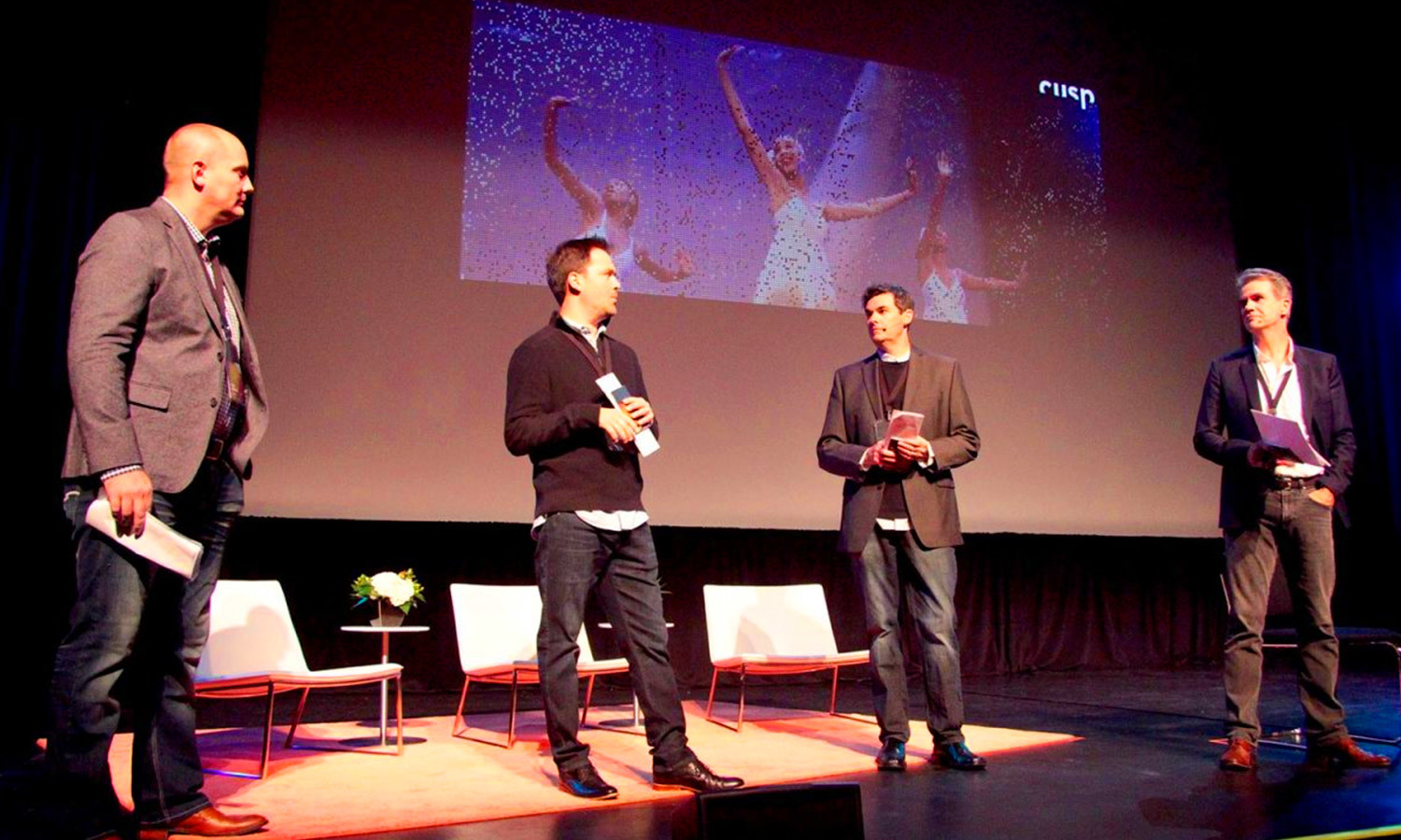 Four men on a stage in a Cusp conference setting while behind them, a large screen displays vibrant dancers.