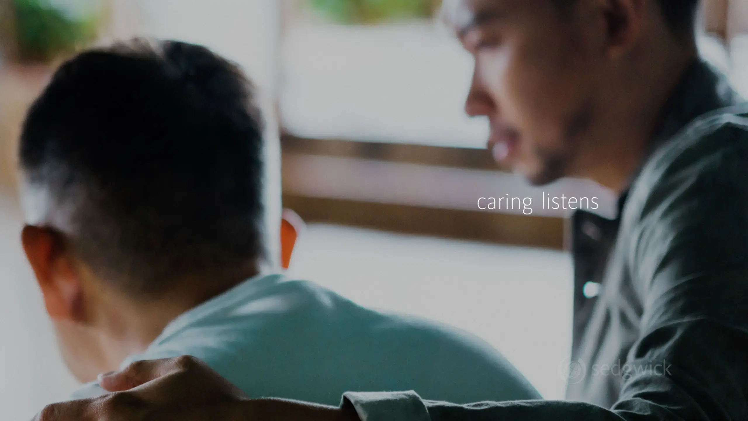 A man places a comforting hand on another's shoulder, conveying empathy. "Caring listens" text emphasizes support and connection in a softly lit room.