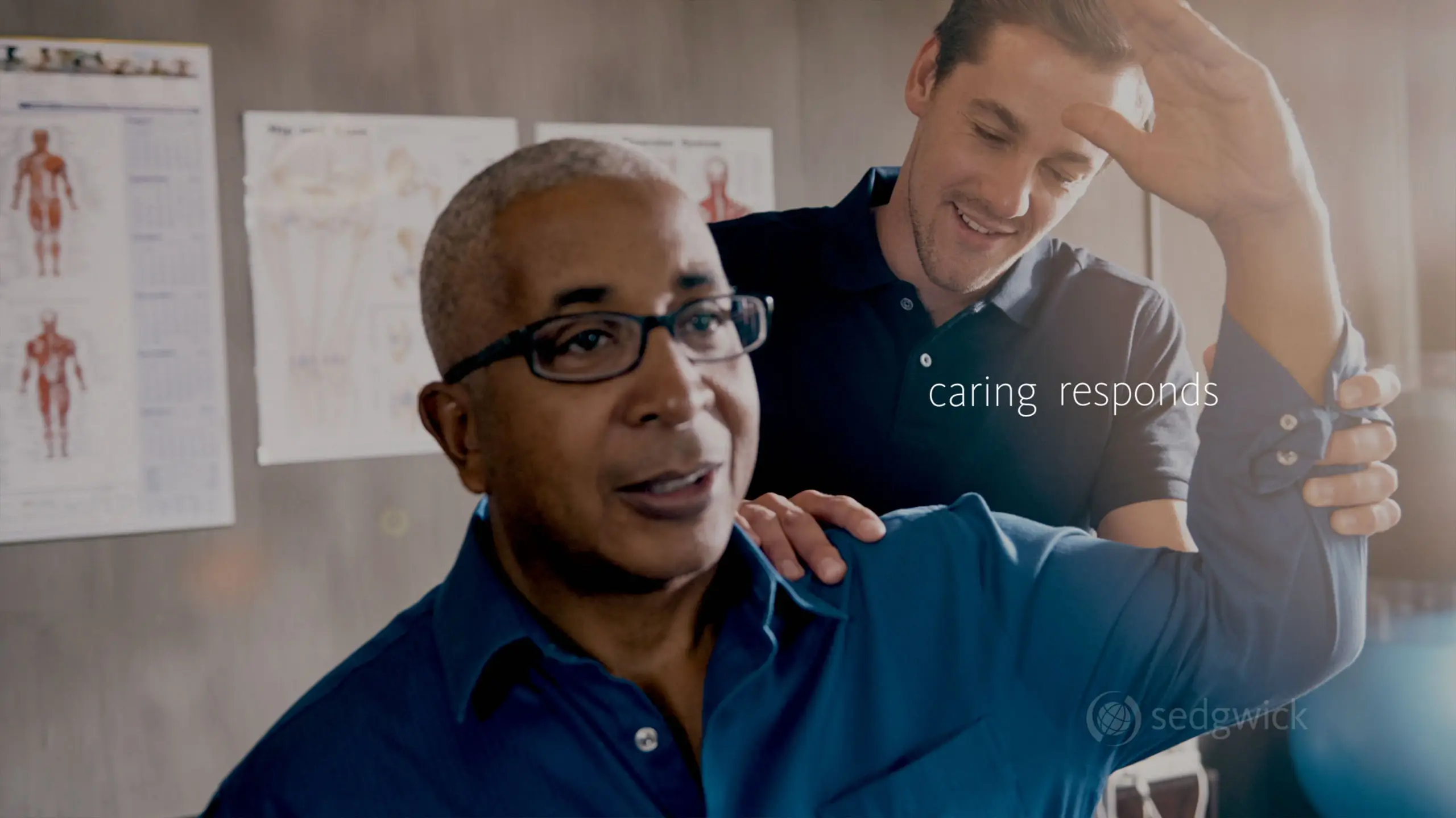 A physical therapist assists an older man in a blue shirt with arm exercises. Medical charts are visible on the wall. The mood is supportive and caring.