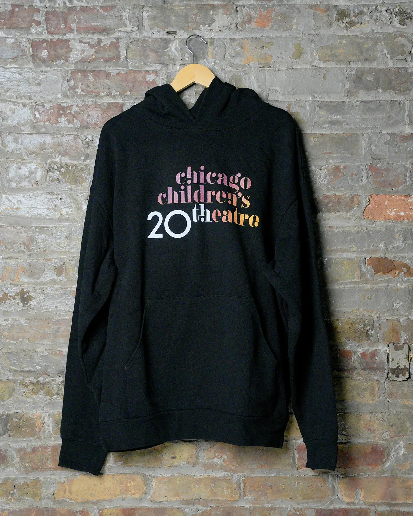 Black hoodie on hanger against brick wall, featuring “Chicago Children’s Theatre 20th” in colorful text.