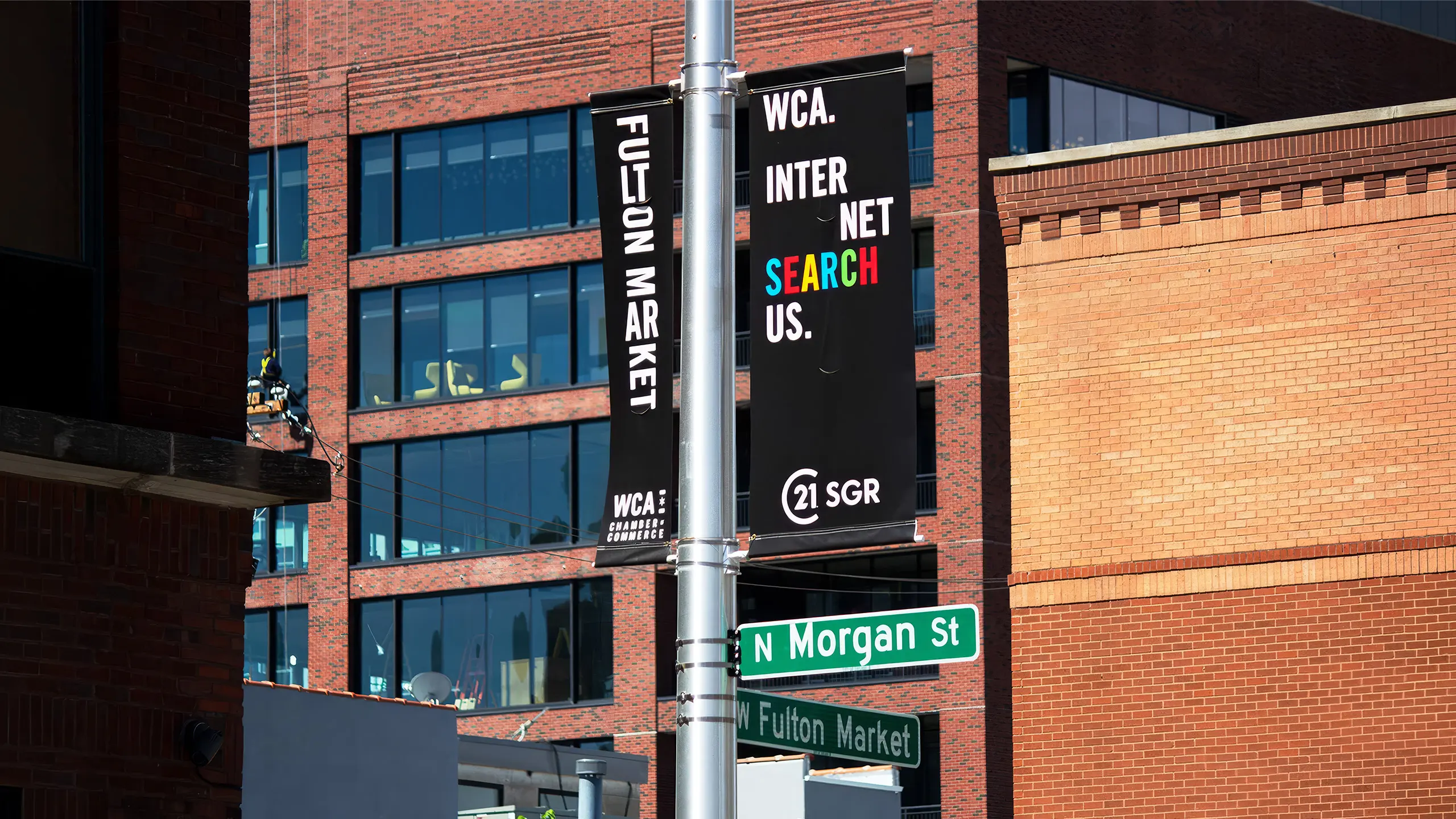 Street signs and banners for Fulton Market and N Morgan St are displayed on a pole in an urban area with brick buildings in the background.